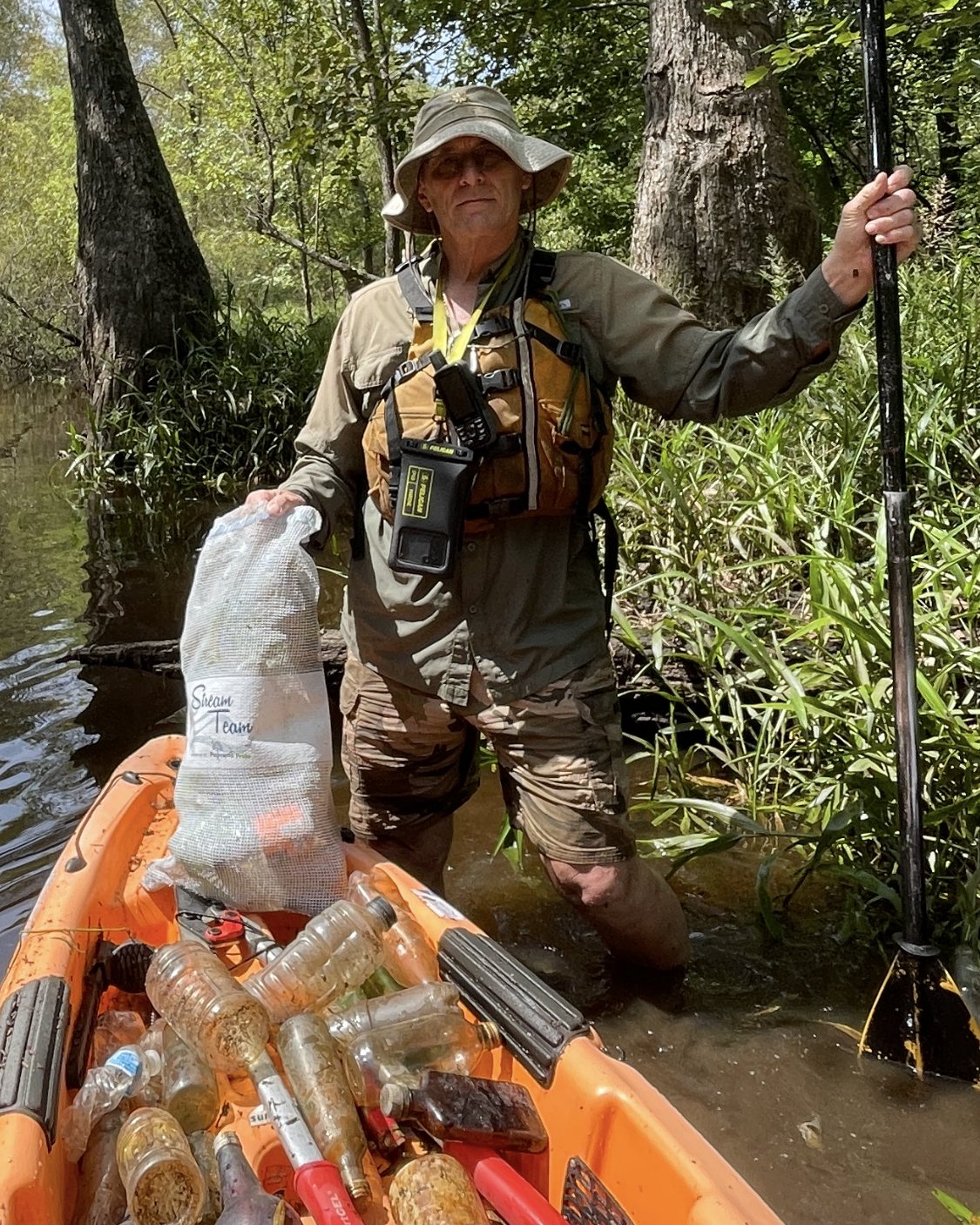 A volunteer in a wide-brim hat and life vest stands in a creek holding a paddle and a mesh bag, beside an orange kayak filled with collected bottles.